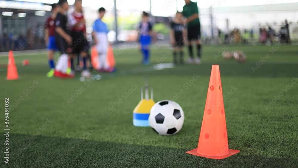 Selective focus to marker cone and soccer training equipment on green artificial turf with blurry kid players training background. Selective focus to marker cone and soccer training equipment on green artificial turf with blurry kid players training background.