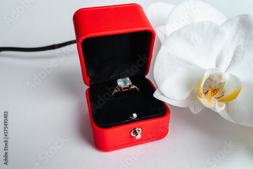 Engagement ring in red gift box on white background. Marry me. Luxury accessory. Place for text. Tropical orchid flowers.