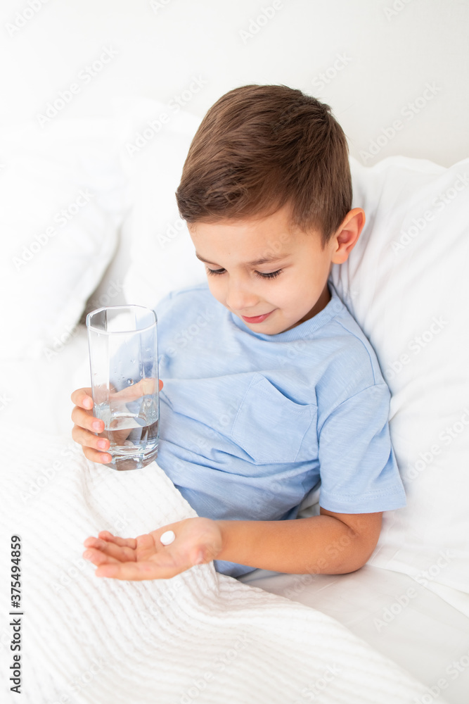 little boy lies sick in white bed and holds a tablet and a glass of water little boy lies sick in white bed and holds a tablet and a glass of water
