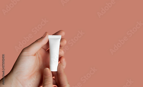 hands with delicate skin and neat nude manicure hold small white clean cosmetic tube on delicate pink background, isolated skin care layout