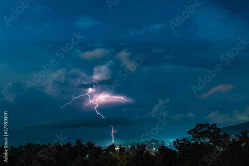 Lightnings, stars and dramatic clouds in the night sky over valley with trees, sea,  city and mountains. View from the top.