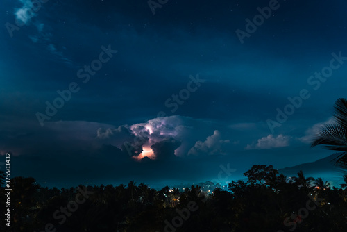Lightnings, stars and dramatic clouds in the night sky over valley with trees, sea,  city and mountains. View from the top.