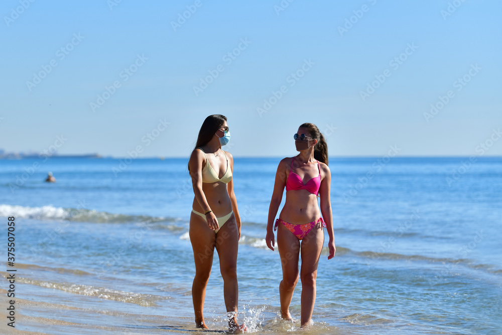 Fototapeta premium Two young women wearing face masks and bikinis talking to each other while walking close to the sea. Safety and new travel concept.