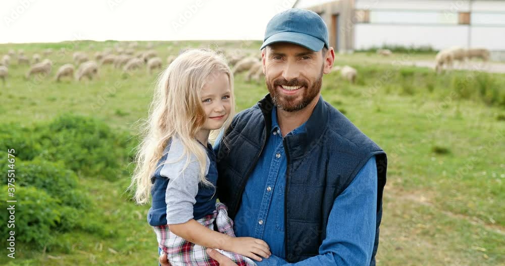 Portrait of Caucasian young handsome father holding on hands cute little daughter and smiling to camera in green field. Sheep flock grazing on background a pasture. Smiled joyful man and small girl. Portrait of Caucasian young handsome father holding on hands cute little daughter and smiling to camera in green field. Sheep flock grazing on background a pasture. Smiled joyful man and small girl.