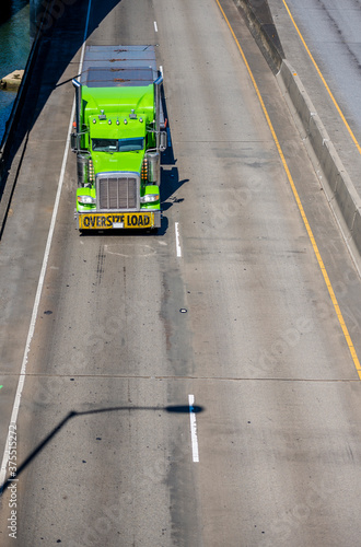Green big rig semi truck with oversize load sign transporting cargo on flat bed semi trailer running on the overpass road along the river