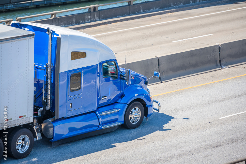 Two color Big rig blue and white semi truck with high cab and dry van semi trailer driving on the divided highway road