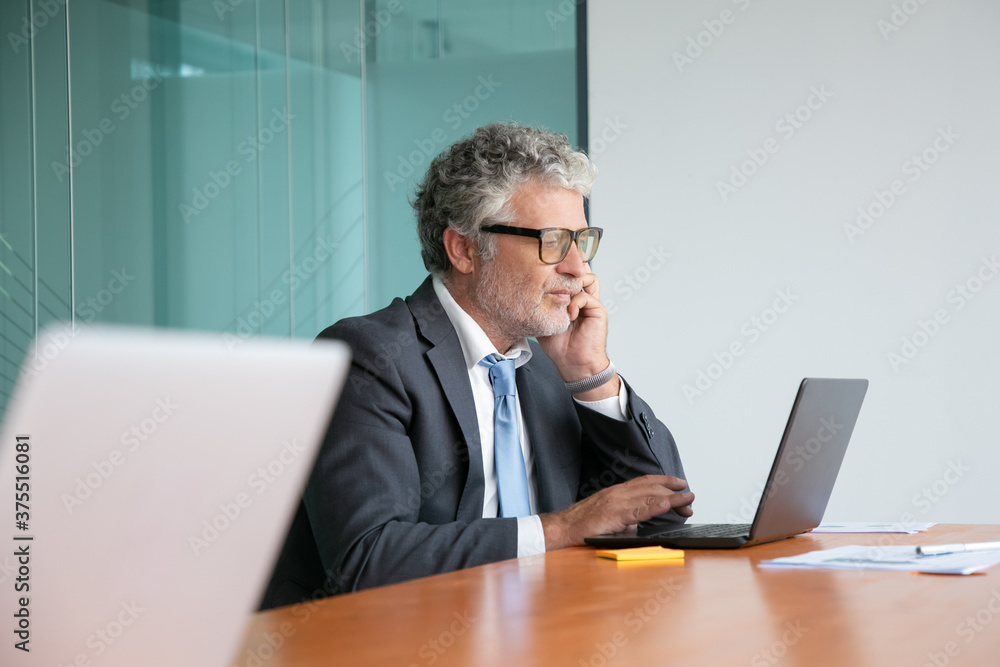 Serious mature professional in suit and glasses talking on cellphone, working at laptop in office, looking at display. Medium shot, side view. Communication or multitasking concept Serious mature professional in suit and glasses talking on cellphone, working at laptop in office, looking at display. Medium shot, side view. Communication or multitasking concept