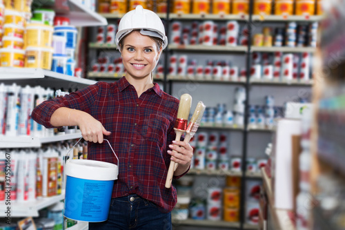 smiling adult woman is standing with paint and brushes for repair house in the store.