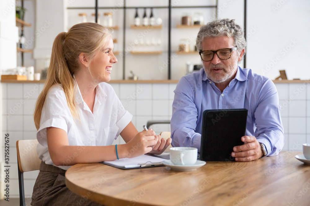 Friendly mature mentor explaining project detail to joyful intern. Business man using tablet and talking to young female colleague. Medium shot. Communication or teamwork concept Friendly mature mentor explaining project detail to joyful intern. Business man using tablet and talking to young female colleague. Medium shot. Communication or teamwork concept