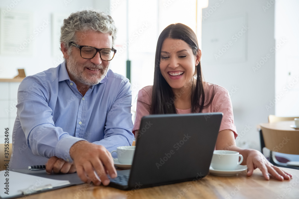 Happy cheerful mature man and young woman sitting at open laptop, looking at display, watching content over cup of coffee and laughing. Medium shot. Digital communication concept Happy cheerful mature man and young woman sitting at open laptop, looking at display, watching content over cup of coffee and laughing. Medium shot. Digital communication concept