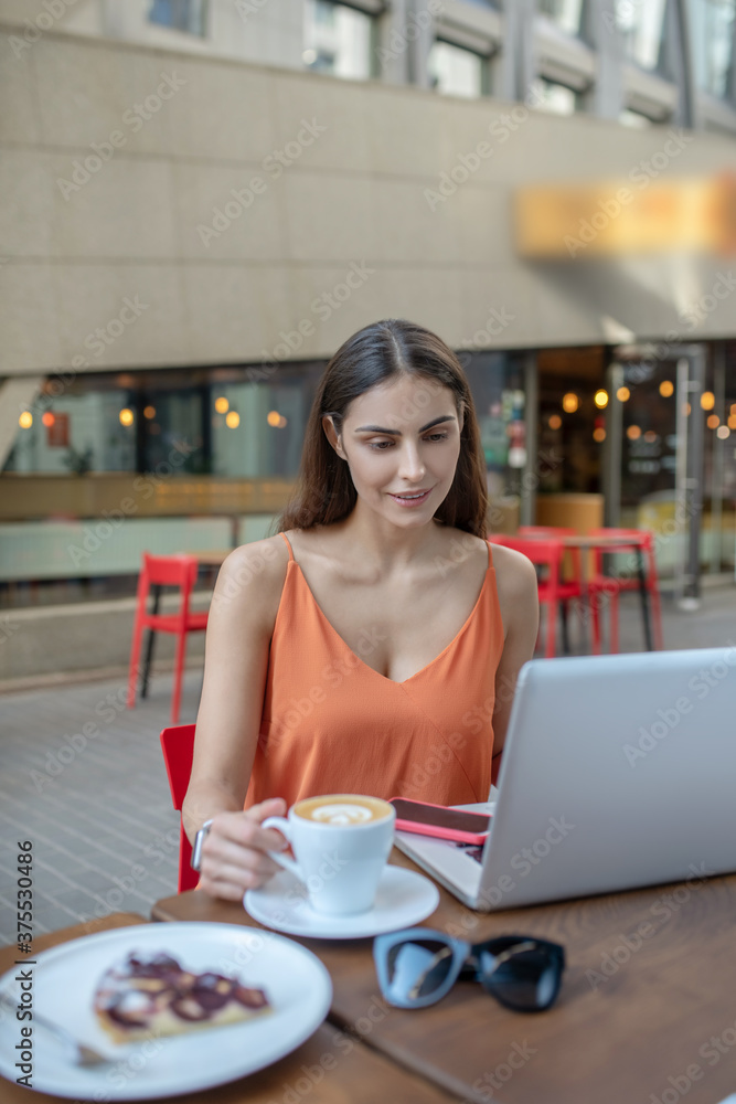 Dark-haired pretty woman having coffee and watching something on a laptop Dark-haired pretty woman having coffee and watching something on a laptop