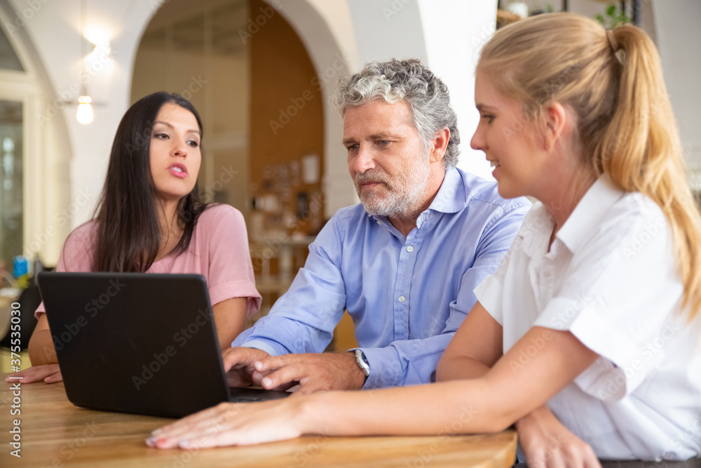 Business team of three sitting at laptop, watching and discussing content. Mature man and young women talking at computer in co-working space. Teamwork or communication concept