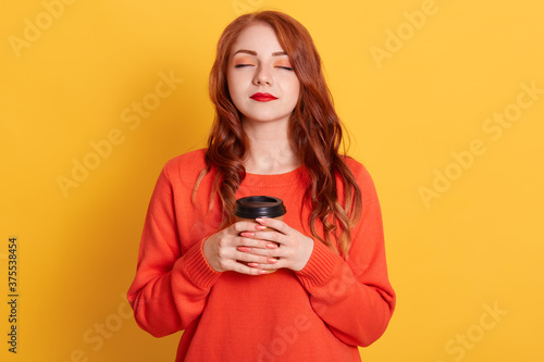 Calm female wearing casual oversize sweater, holding takeaway coffee in both hands, enjoying hot beverage with closed eyes, winsome girl with against yellow background.