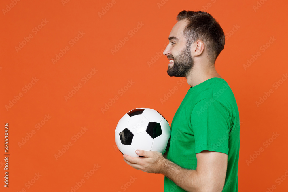 Side view smiling young man football fan in basic green t-shirt cheer up support favorite team with soccer ball isolated on orange background studio portrait. People sport leisure lifestyle concept. Side view smiling young man football fan in basic green t-shirt cheer up support favorite team with soccer ball isolated on orange background studio portrait. People sport leisure lifestyle concept.