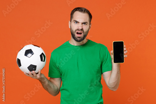 Puzzled young man football fan in green t-shirt cheer up support favorite team with soccer ball hold mobile phone with blank empty screen isolated on orange background. People sport leisure concept.
