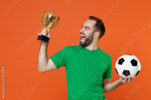 Overjoyed young man football fan in green t-shirt cheer up support favorite team with soccer ball hold golden cup isolated on orange background studio portrait. People sport leisure lifestyle concept.