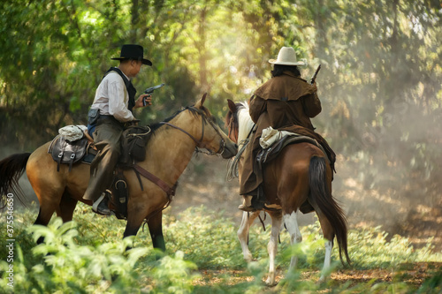 Two cowboy is on the horse, standing in the forest and holding a gun.