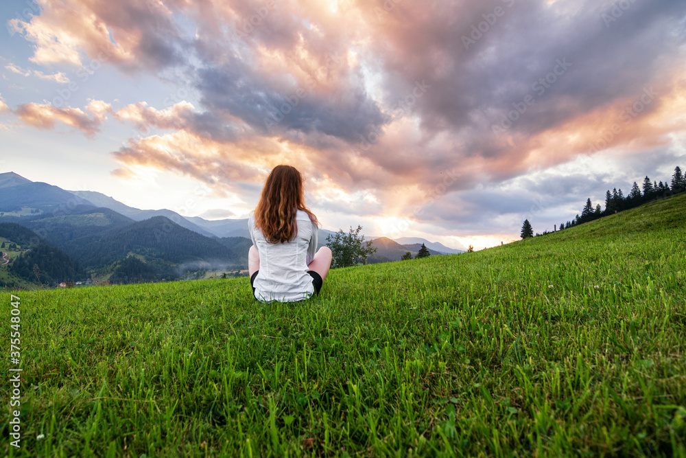 Girl in a white shirt with red hair sitting on the lawn in the mountains. Green grass and sunset in the Ukrainian Carpathians. Girl in a white shirt with red hair sitting on the lawn in the mountains. Green grass and sunset in the Ukrainian Carpathians.