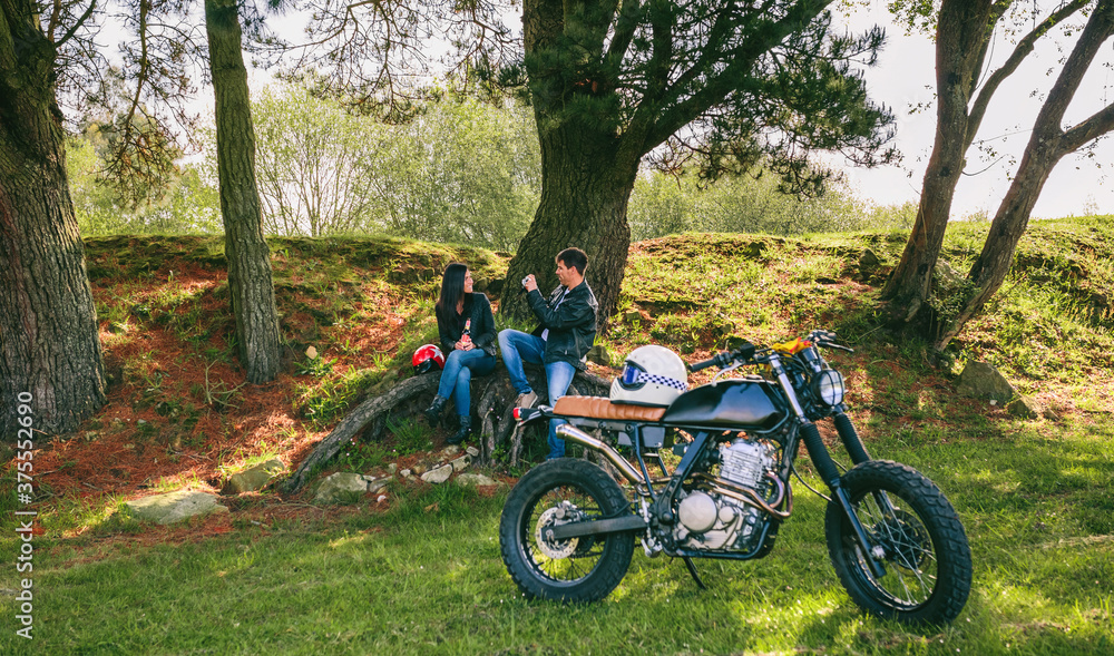 Fototapeta premium Young couple resting from a motorcycle trip taking picture outdoors with motorbike in foreground. Selective focus on couple in the background