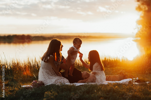 Happy family, mom, son and two girls sitting on the shore of river at summer picnic at sunset. The concept of organic farming and healthy lifestyle, healthy food, happiness and joy. Copy space.