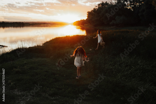 Two little sisters walk on the green grass on the river beach on a summer sunset. Cute little girls in white dresses. 