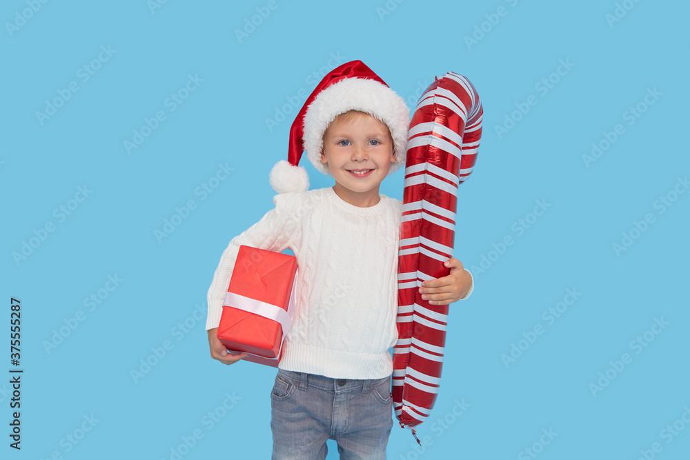 Obraz premium Portrait of a satisfied little child boy in christmas Santa hat. laughing isolated over blue background. Holds a gift box and big candy cane. Preparing for the New Year holidays