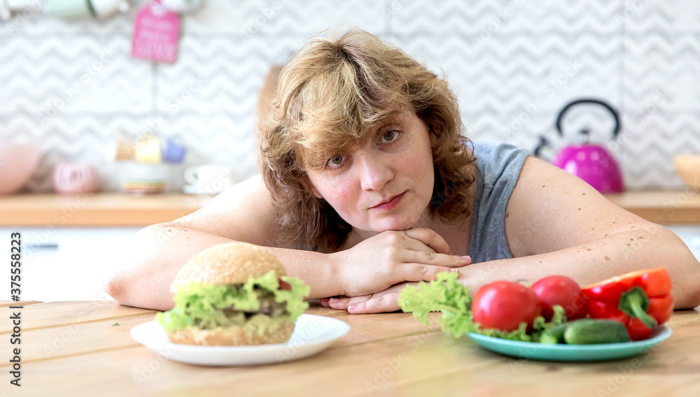 A curvy woman is sitting at a table in the kitchen in her apartment. There's a vegetable salad and a Burger on the table.
