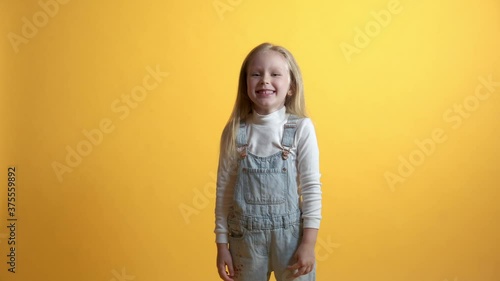 The blonde girl laughs and points a finger to the camera. Contempt, ridicule concept. Yellow background. Girl in denim overalls casual wear.