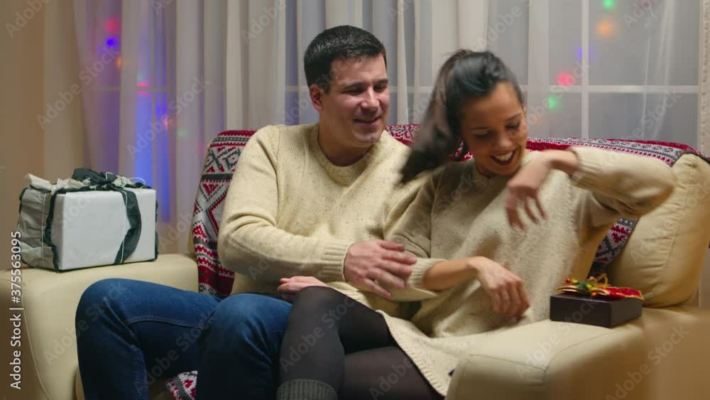 Caucasian young couple on christmas day sitting on couch and giving gifts to each other. Receiving Xmas surprise from loved one. Giving happy romantic present gift box to significant other in new year Caucasian young couple on christmas day sitting on couch and giving gifts to each other. Receiving Xmas surprise from loved one. Giving happy romantic present gift box to significant other in new year
