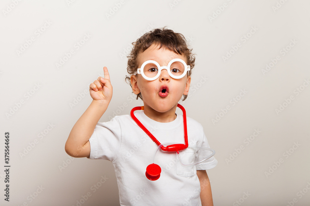 funny curly kid in glasses with a stethoscope on his neck plays a doctor. Close-up of a child, a boy showing surprise and holding a finger up. idea. funny curly kid in glasses with a stethoscope on his neck plays a doctor. Close-up of a child, a boy showing surprise and holding a finger up. idea.
