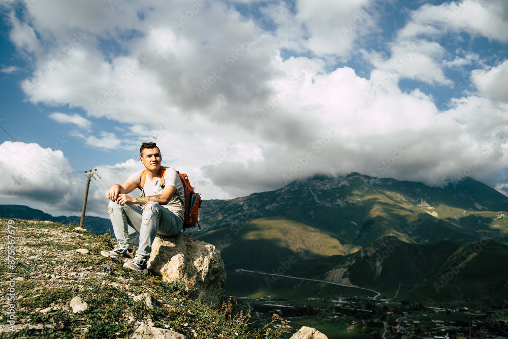 Naklejka premium Young man sitting on hill. Dreamy male sitting on altitude and looking at Peaks of magnificent rocks located against bright cloudy sky