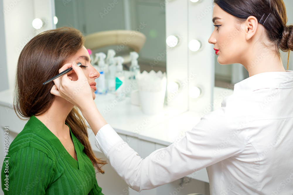 Makeup artist applies eyeshadow powder on eyes of young caucasian woman by brush tool in beauty salon Makeup artist applies eyeshadow powder on eyes of young caucasian woman by brush tool in beauty salon