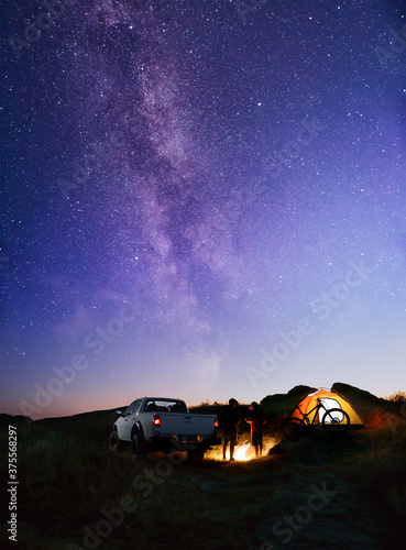 Friends near Bonfire, Pickup Truck, Tent and Bike in the Mountains under Night Sky with Milky Way. Adventure and Travel
