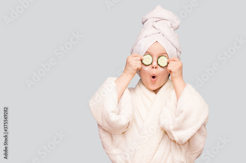 Funny little girl in white coat and towel with mask for skin face and cucumbers on eyes isolated on white background