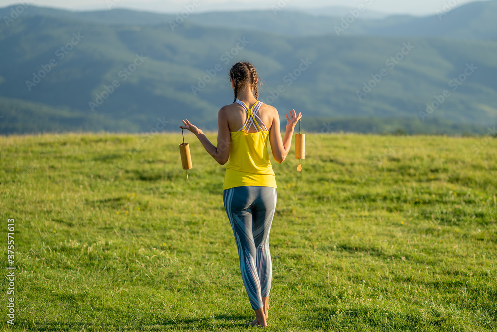 Obraz premium Full body shot of a slim caucasian woman performing guided meditation through music using koshi chimes on a green meadow on a beautiful summer morning. Beautiful girl holding wind chimes in her hands.