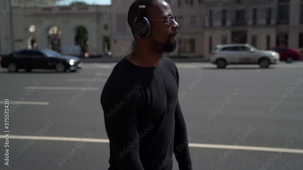 A stylish African American man walks through the city on a Sunny day next to the road and listens to his favorite music with headphones A stylish African American man walks through the city on a Sunny day next to the road and listens to his favorite music with headphones