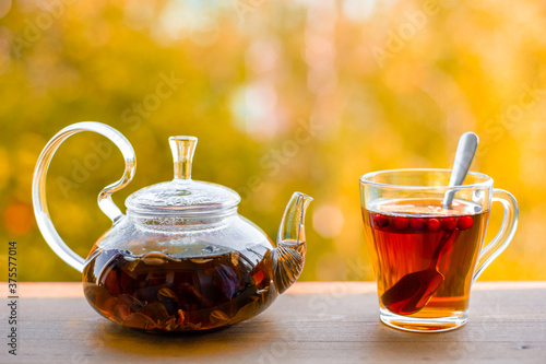 Warming autumn tea outdoors. Glass cup and kettle with herbal tea and berries on a wooden table against the background of yellow trees.