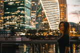 A young woman stands at night in front of glowing glass skyscrapers in Moscow. High ISO, grainy and noisy images.
