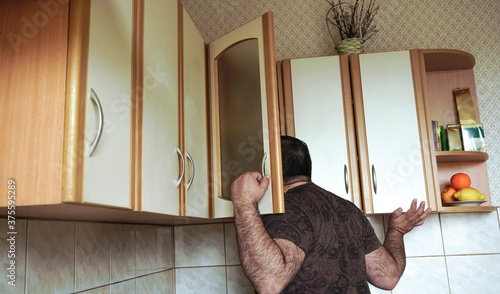 Obraz na plátně Young man in shirt peeking  into kitchen cupboard at home, viewed from the back side