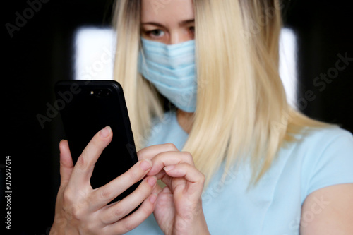 Blonde woman in face mask using smartphone, mobile phone in female hands closeup. Concept of safety work in office during coronavirus pandemic
