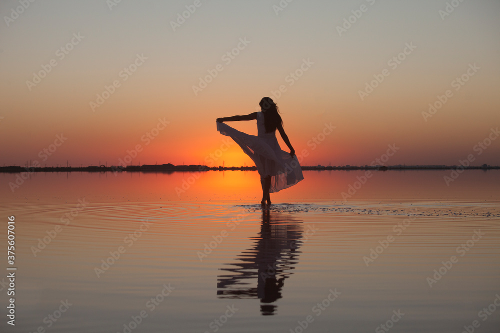 Woman walking on the water silhouette in a beautiful dress on the lake in the evening light at sunset