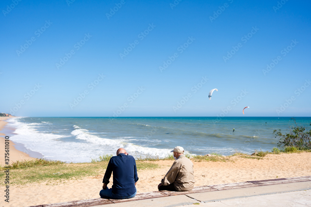 Fototapeta premium Back view of old men sitting on the bench near ocean, kiteboarding or kitesurfing on sunny outdoors background.