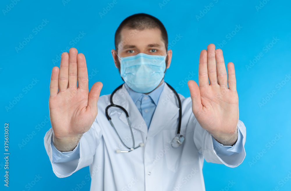 Doctor in protective mask showing stop gesture on light blue background. Prevent spreading of coronavirus Doctor in protective mask showing stop gesture on light blue background. Prevent spreading of coronavirus