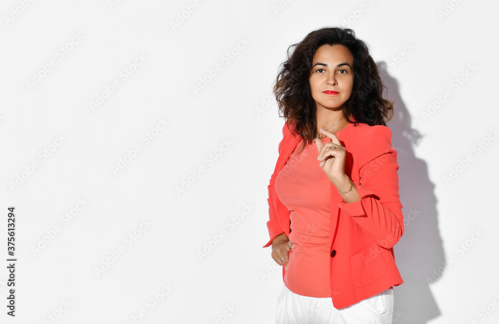 Stylish curly brunette woman model in elegant coral jacket and white pants stands at copy space holding hand on her waist and finger up Stylish curly brunette woman model in elegant coral jacket and white pants stands at copy space holding hand on her waist and finger up