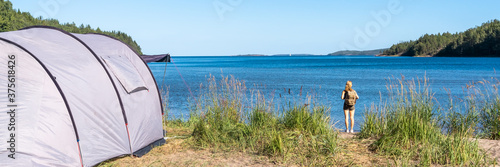 A slim athletic girl with a backpack on the shore of the sea bay looks at the horizon. Young sporty woman traveller  is engaged in hiking at sunny day. Tourist equipment for trekking. Camping tents.