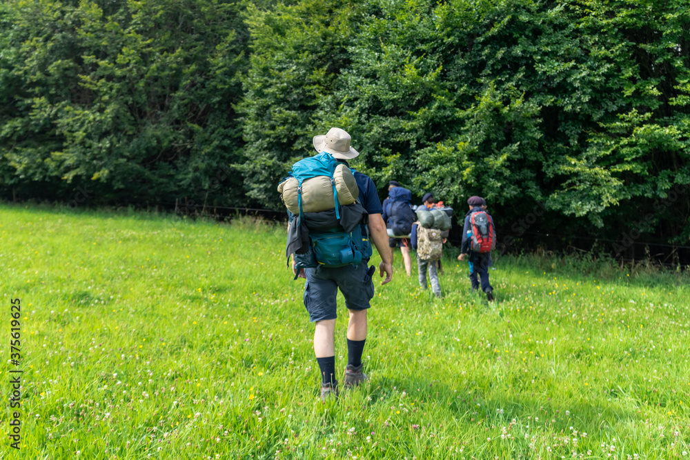 Scouts or tourists go with big backpacks on the green forest trail on sunny summer day. Active healthy lifestyle people with rucksacks. Hiking Trekking Scouting. Green forest background.