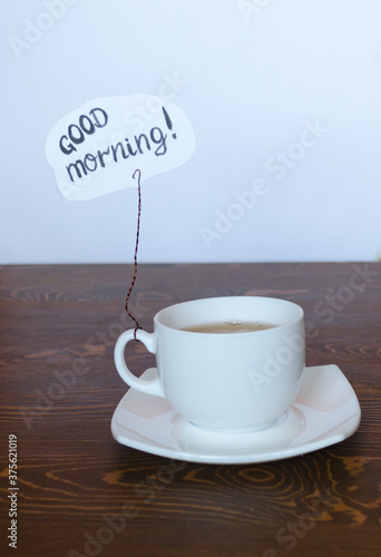 Cup with tea close-up. A sign with the inscription good morning. Inspiration to start a new day. White background and wood texture. White dishes for a drink on the table.