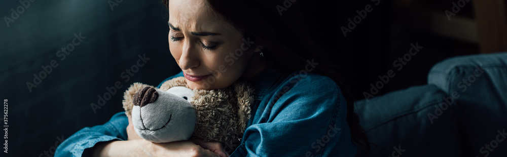 panoramic crop of sad brunette woman with closed eyes holding teddy bear in living room panoramic crop of sad brunette woman with closed eyes holding teddy bear in living room