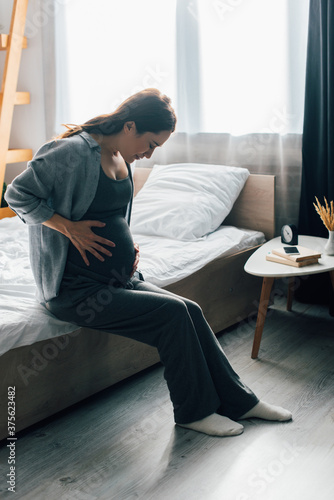 brunette pregnant woman suffering from pain while sitting on bed