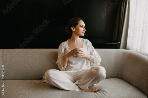 Young woman with calm mood, sitting on a sofa, wearing white silk sleepwear and holding a cup of coffee.